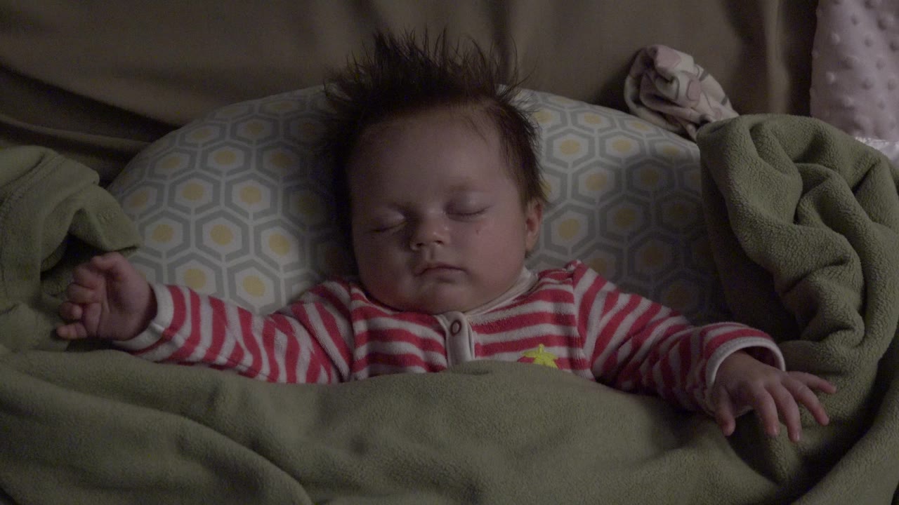A close indoor view in Westminster, Colorado shows the sleeping baby centered between patterned bedding and a folded green blanket at the right edge.
