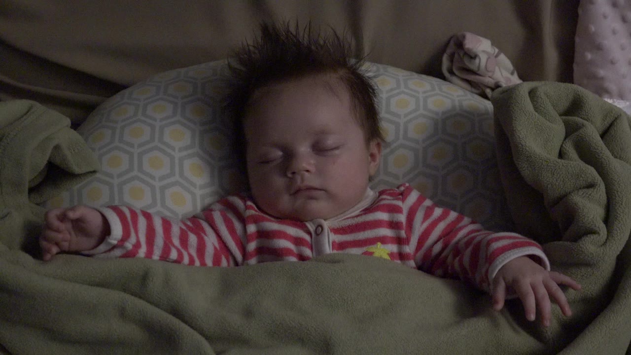 At 0:00, a sleeping baby with dark tousled hair lies under a beige-green blanket on patterned bedding in Westminster, Colorado.