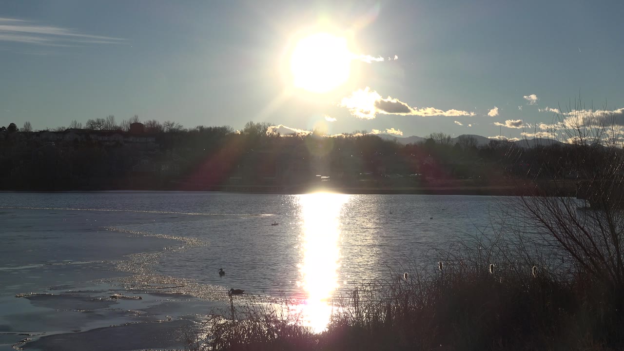 A bright sun sits above the far bank of the Westminster, Colorado lake, with the water reflecting white light below it, dark trees on the horizon, and no visible people.