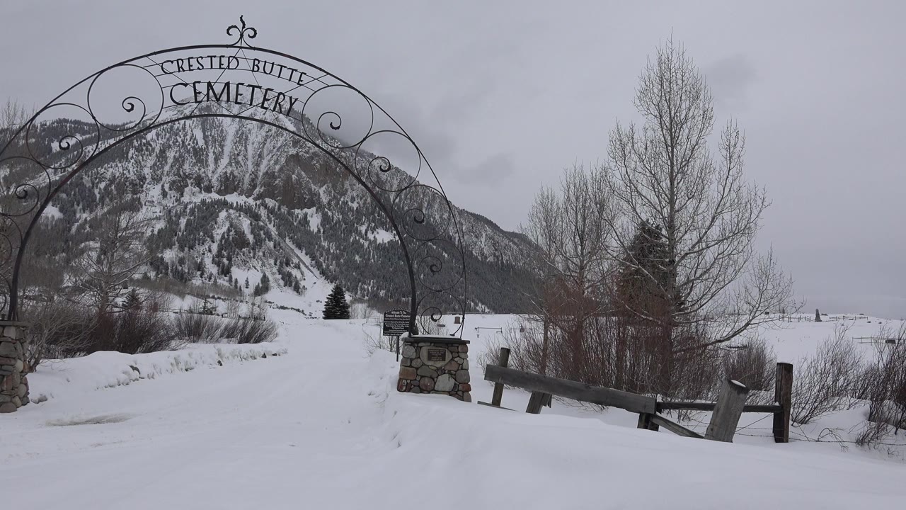 Snowy cemetery entrance with black iron arch and mountain behind it.