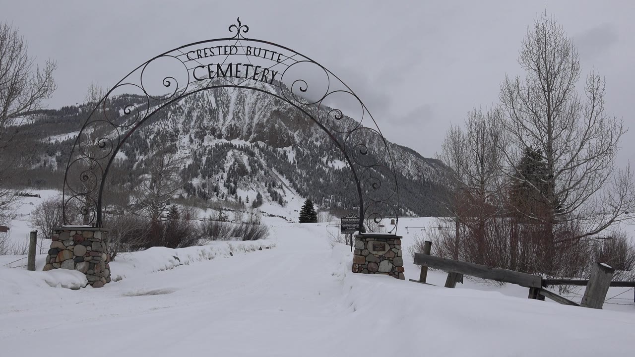Snowy cemetery entrance with iron arch and mountain behind it.