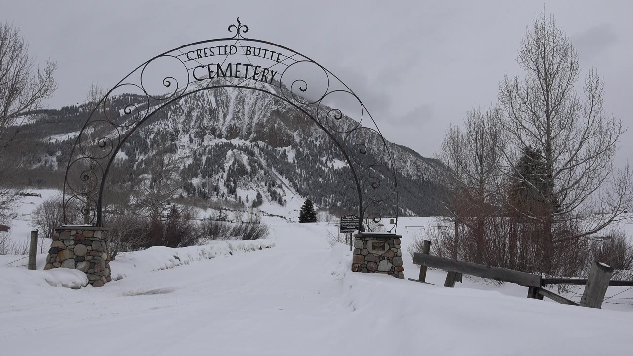 A near-centered view of the Crested Butte Cemetery arch shows the dark ironwork against gray sky, with snowy ground, leafless trees, and the mountain slope visible through the gate, with no visible people.