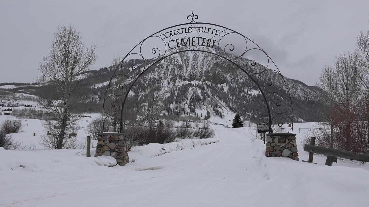 Crested Butte Cemetery arch remains centered over the white path, with the left bare tree and snowy mountain slope visible through the ironwork, and no visible people.