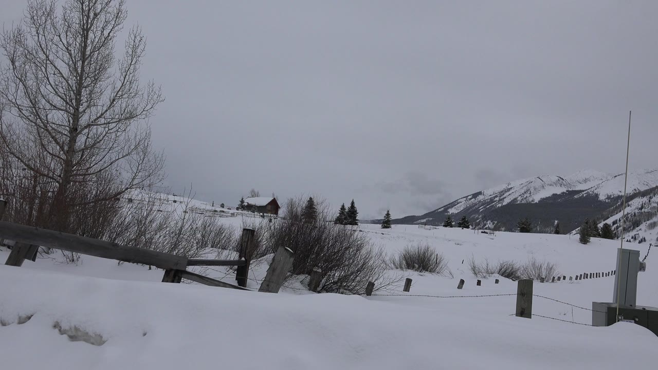 Opening frame of a snowy Crested Butte hillside with a low wooden fence in the foreground, a small house on the slope, distant mountains, gray dusk sky, and no visible people.