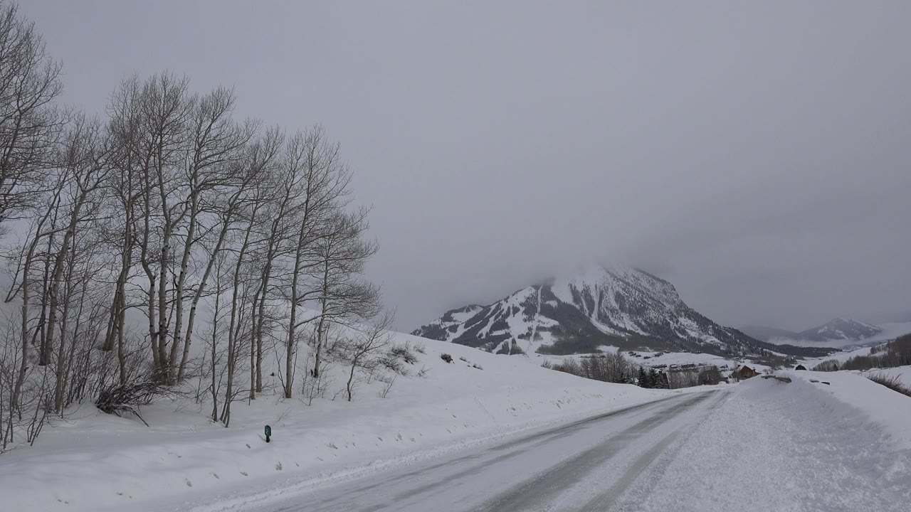Snow-covered road in Crested Butte, Colorado leading toward dark mountain peaks, with bare trees on the left and no visible people.