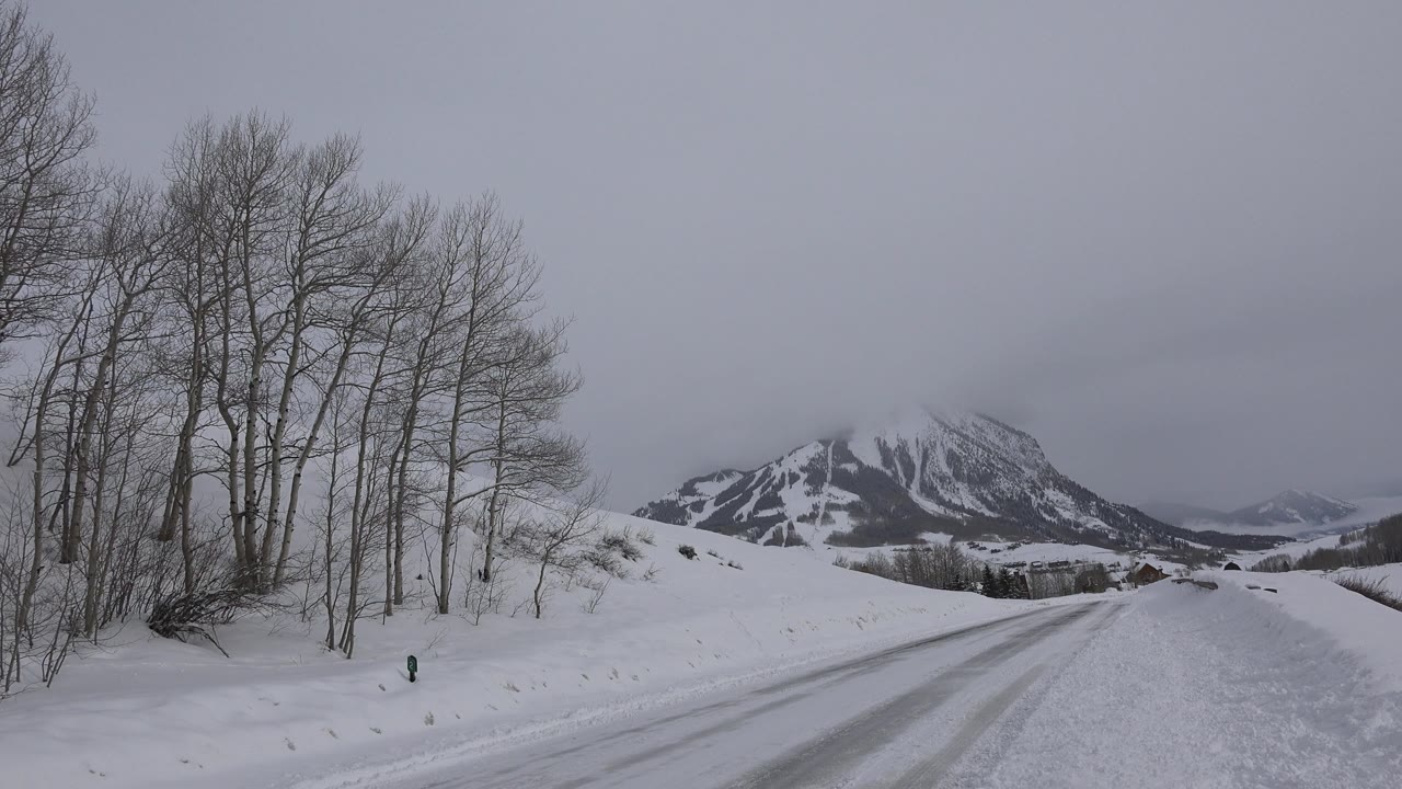 A snowy Crested Butte road fills the foreground while leafless trees line the left side and a cloud-covered mountain ridge sits ahead, with no visible people.