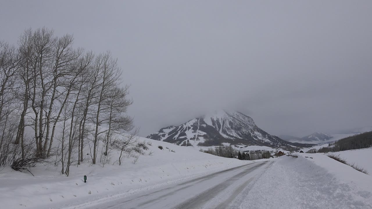 Snow-covered road in Crested Butte, Colorado leading toward snow-streaked mountains, with bare trees on the left and no visible people.