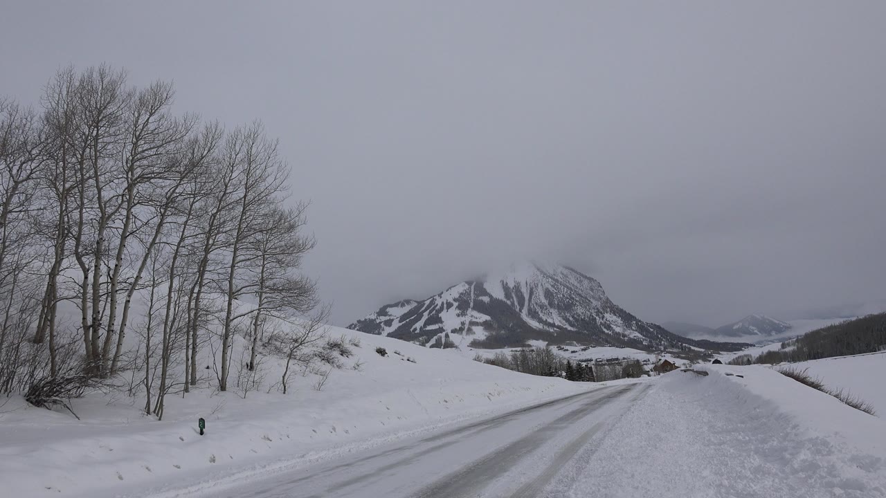 Crested Butte winter road with leafless trees close to the left edge, snowy banks beside the road, distant buildings near the mountain base, and no visible people.