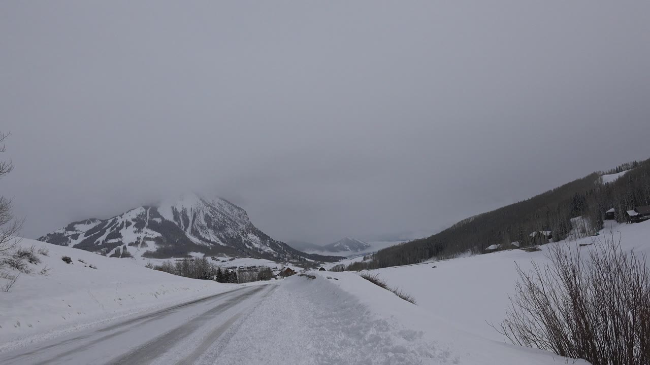 Snowy road leading toward mountains under a gray sky.