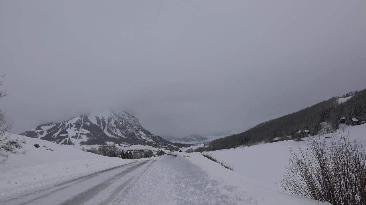 The road remains empty as foreground shrubs and snowbanks frame the view toward the Crested Butte mountain valley, with no visible people.