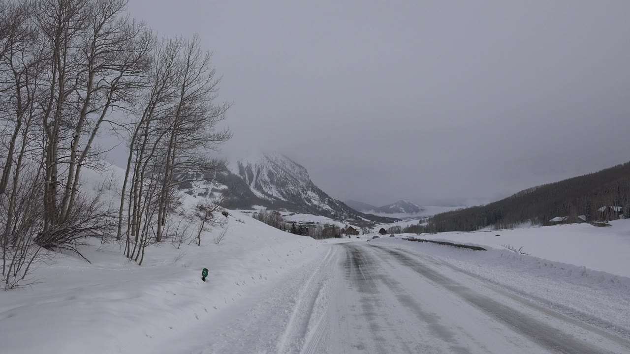 A second view down the snowy Crested Butte road shows tire tracks, a stand of bare roadside trees, muted hillsides, and no visible people.