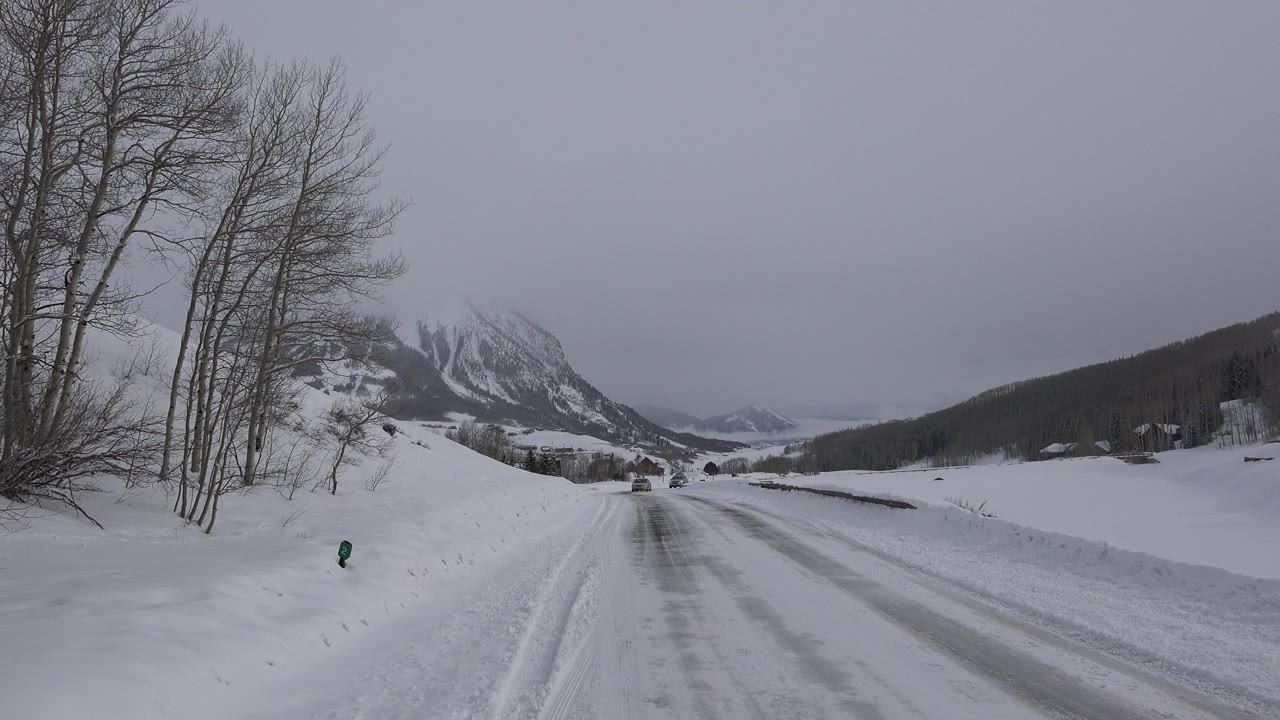 Snow-covered road curving past bare trees toward distant mountains.