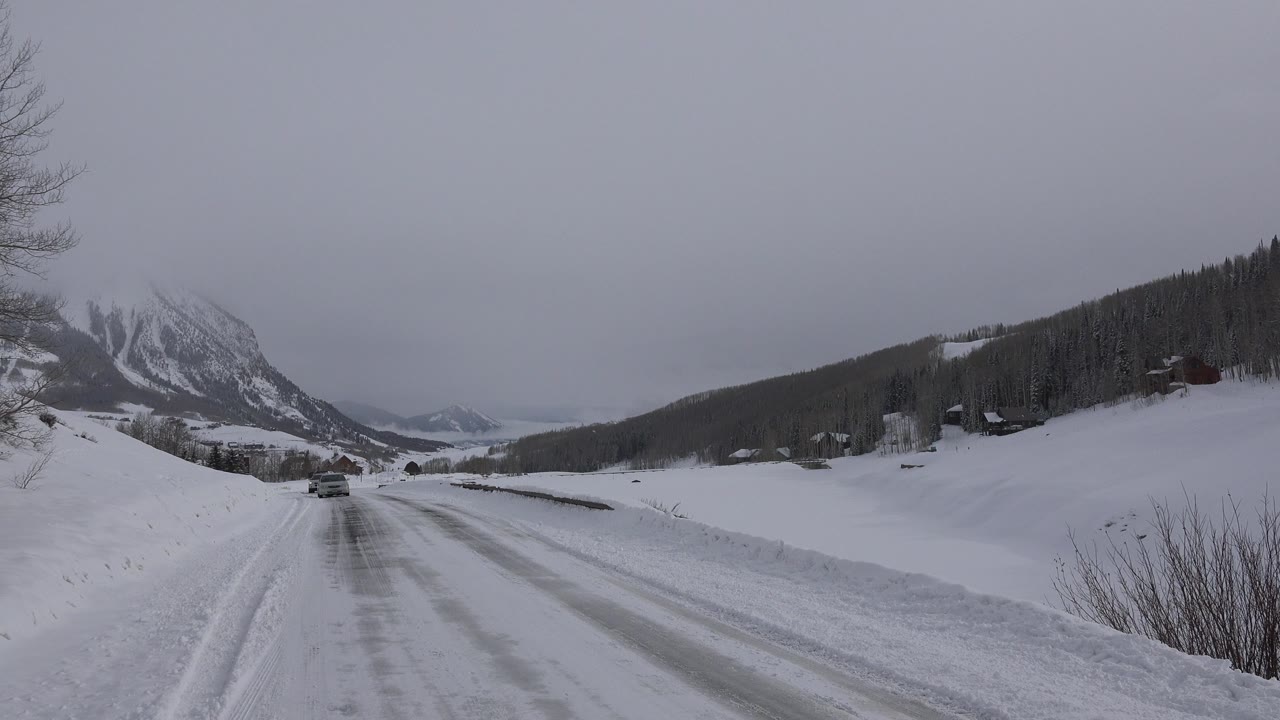 Opening frame of a snowy road in Crested Butte, Colorado, with a vehicle on icy pavement, snowbanks, distant mountains, and no visible people.