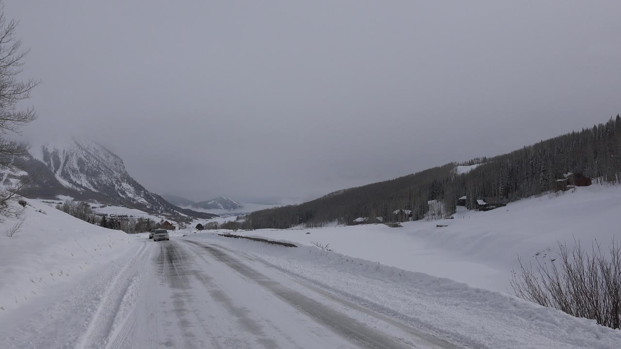 Second frame shows the same Crested Butte winter road with the vehicle slightly nearer, snow piled along both sides, dark trees on the hillside, and no visible people.