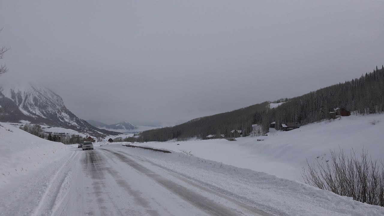 Vehicle on a snowy mountain road with cabins on a forested slope.