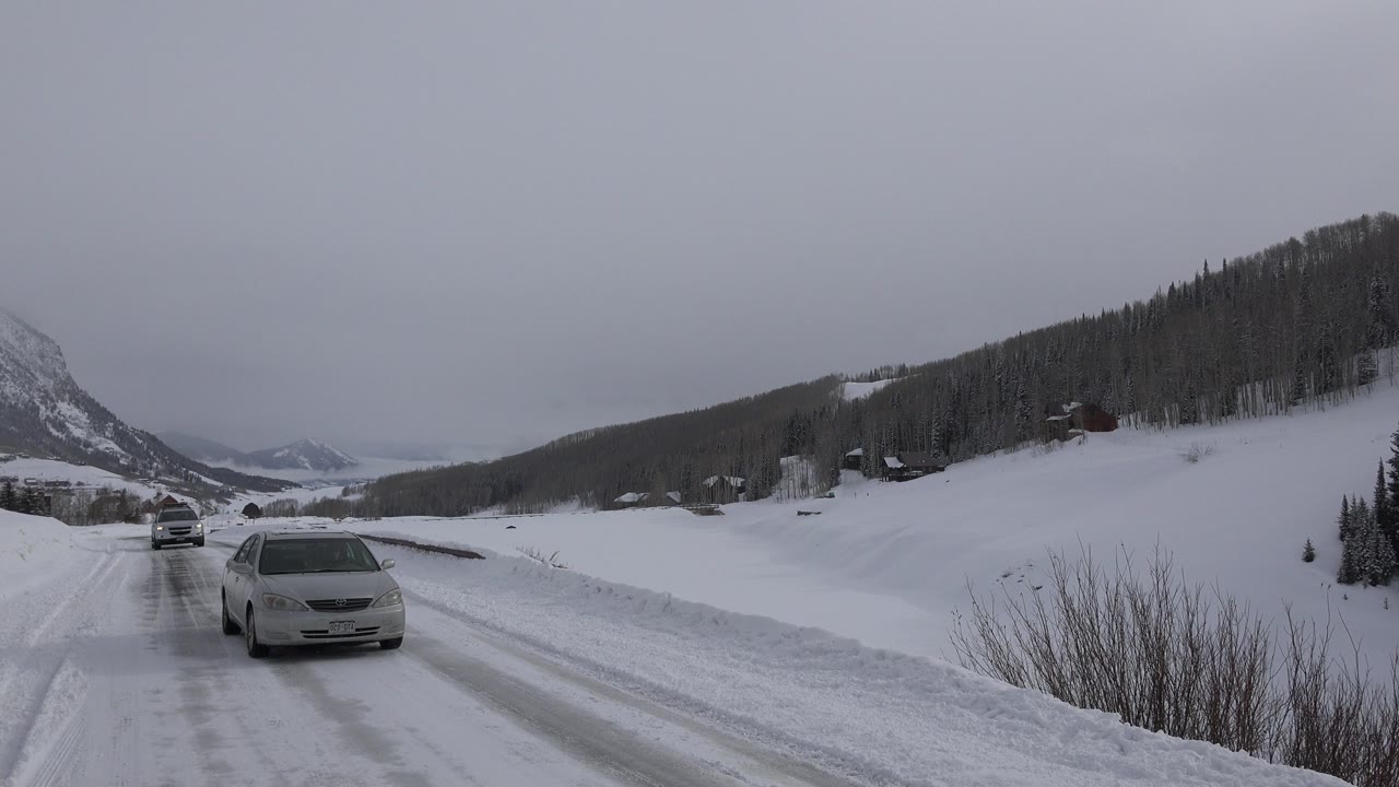 The car moves closer along the snowy Crested Butte road while the curving shoulder, dark hillside trees, and overcast mountain background remain visible, with no visible people.