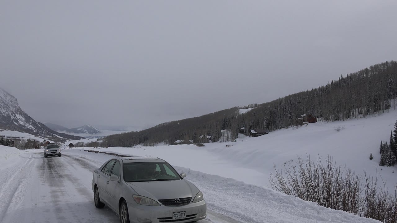 A light-colored sedan passes close to the roadside camera on a snowy Crested Butte roadway, with white hills, dark trees, and no visible people in the scene.