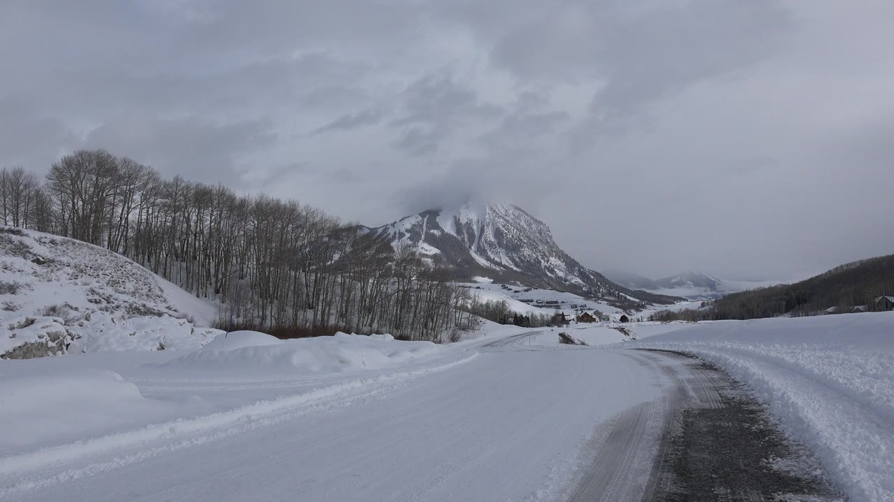 Snowy road in Crested Butte, Colorado curving between high snowbanks toward a distant icy mountain, with bare trees on the left and no visible people.