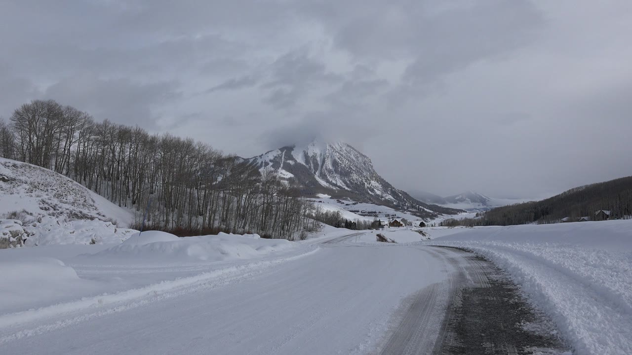 Snowy road curving toward mountains under gray clouds.