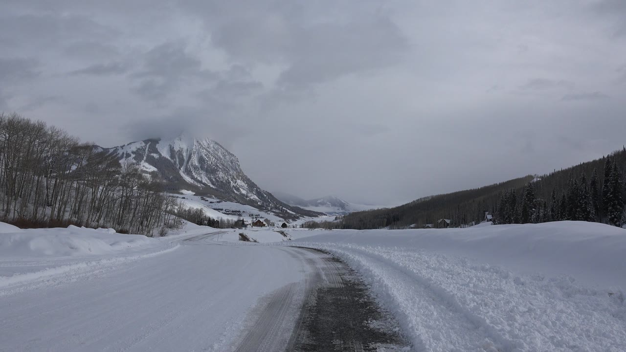 Snow-covered road leading toward mountains under gray clouds.
