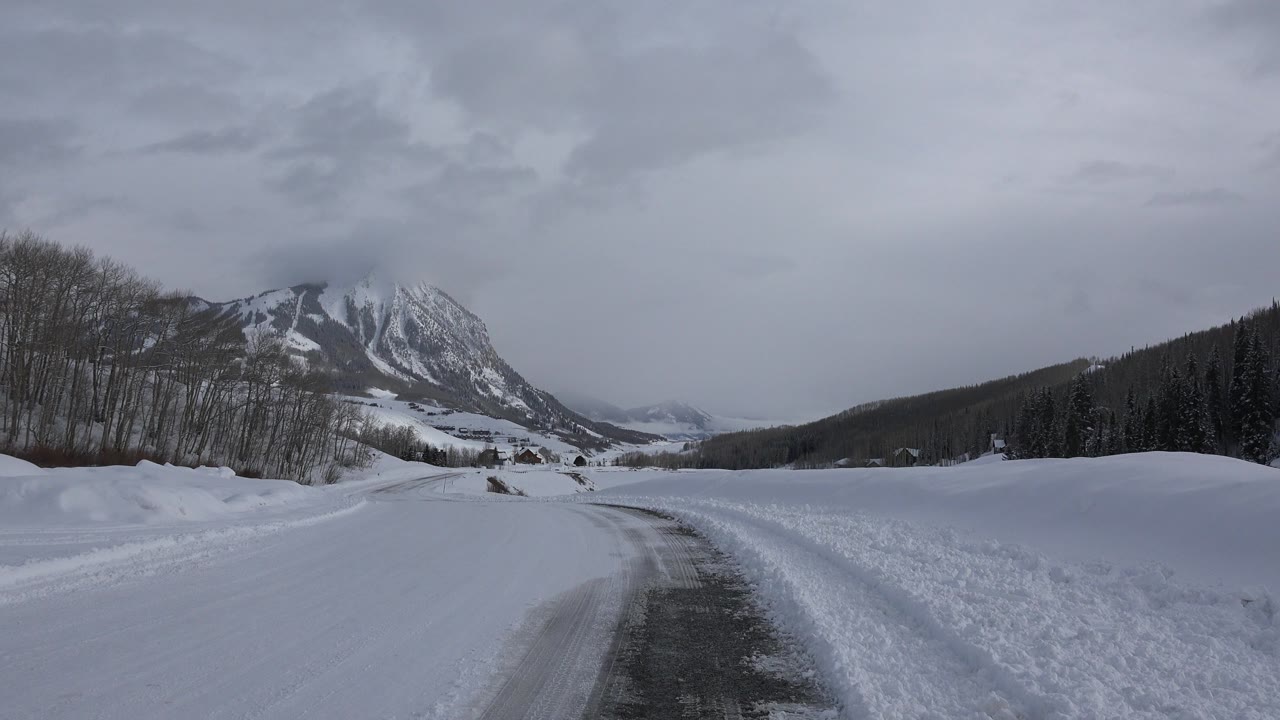 At 0:01, the icy road sits slightly right of center in Crested Butte, Colorado, with deep snow on both sides, a dark tree line, distant buildings, and no visible people.