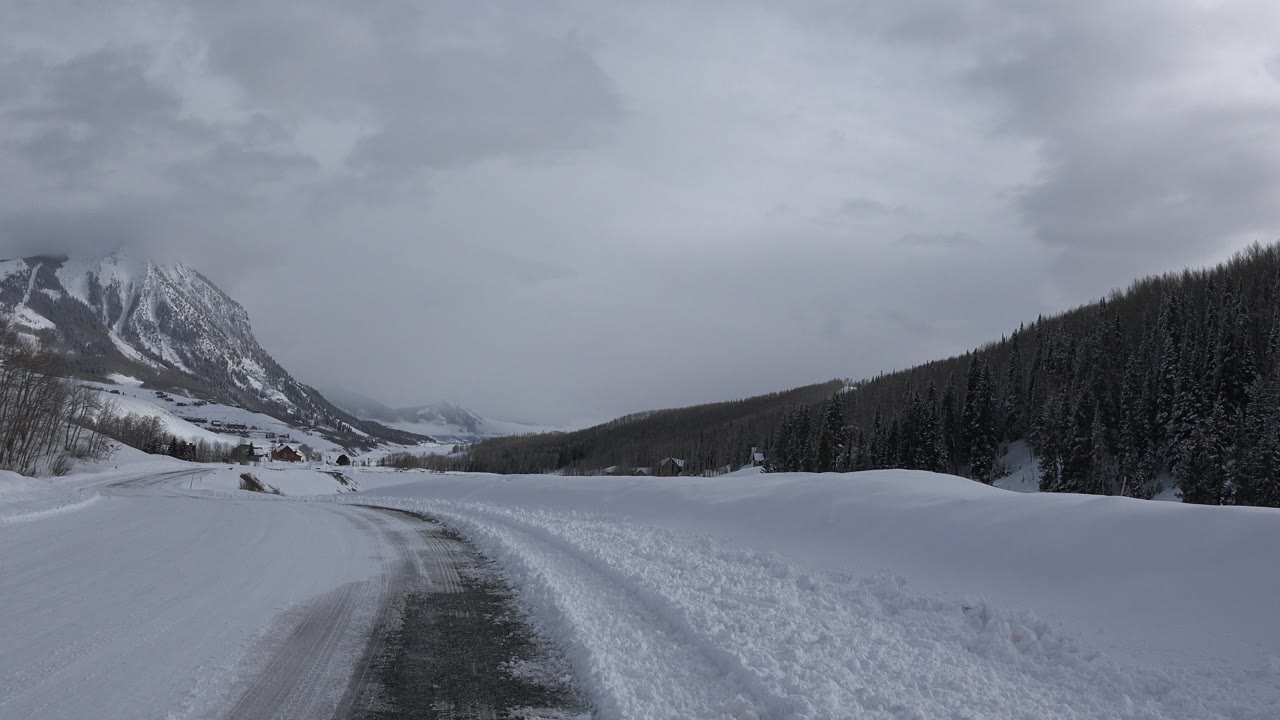 Snow-covered road beside a mountain valley and evergreen slope.