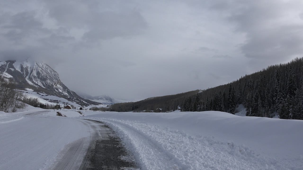 At 0:01 in Crested Butte, Colorado, the icy road curves through white snowbanks with a rocky mountain on the left and dark evergreens on the right, with no visible people.