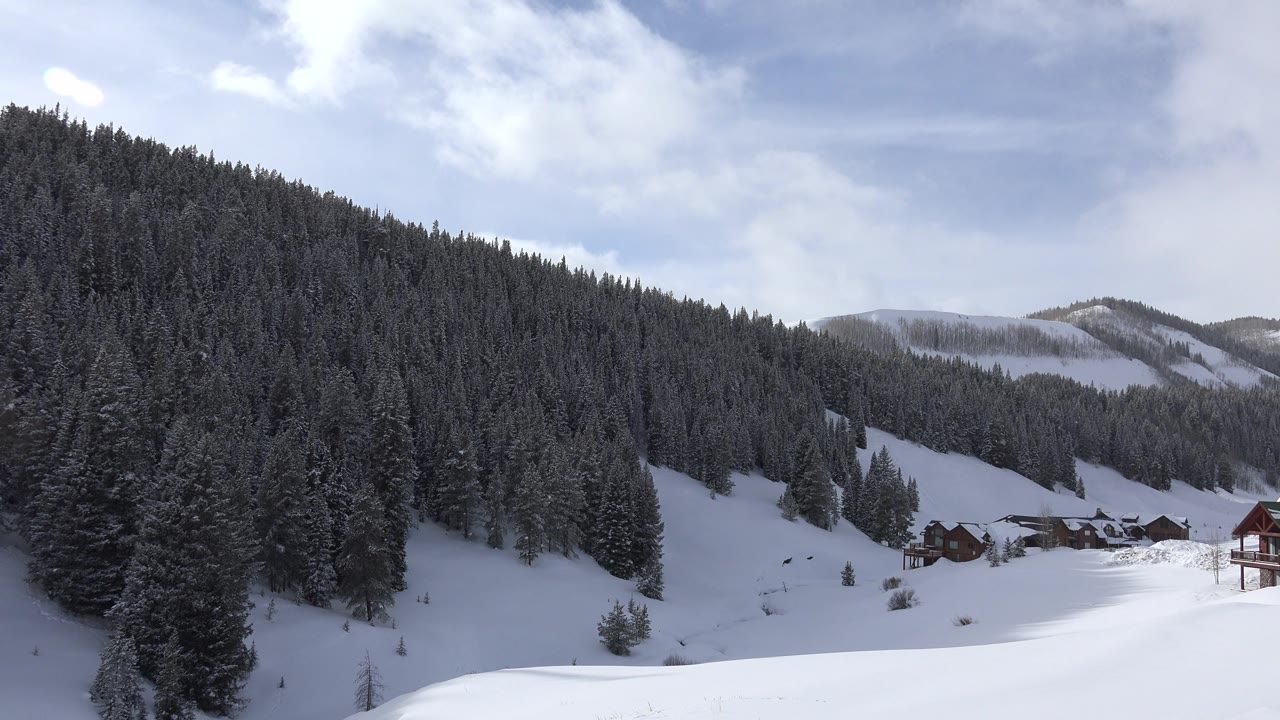 Wide snowy valley edge with forest, cabins, and mountains under clouds.