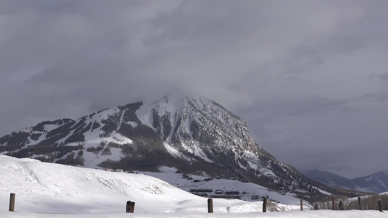 Opening view of a snow-covered mountain landscape in Crested Butte, Colorado, with dark forest patches, gray clouds, foreground fence posts, and no visible people.