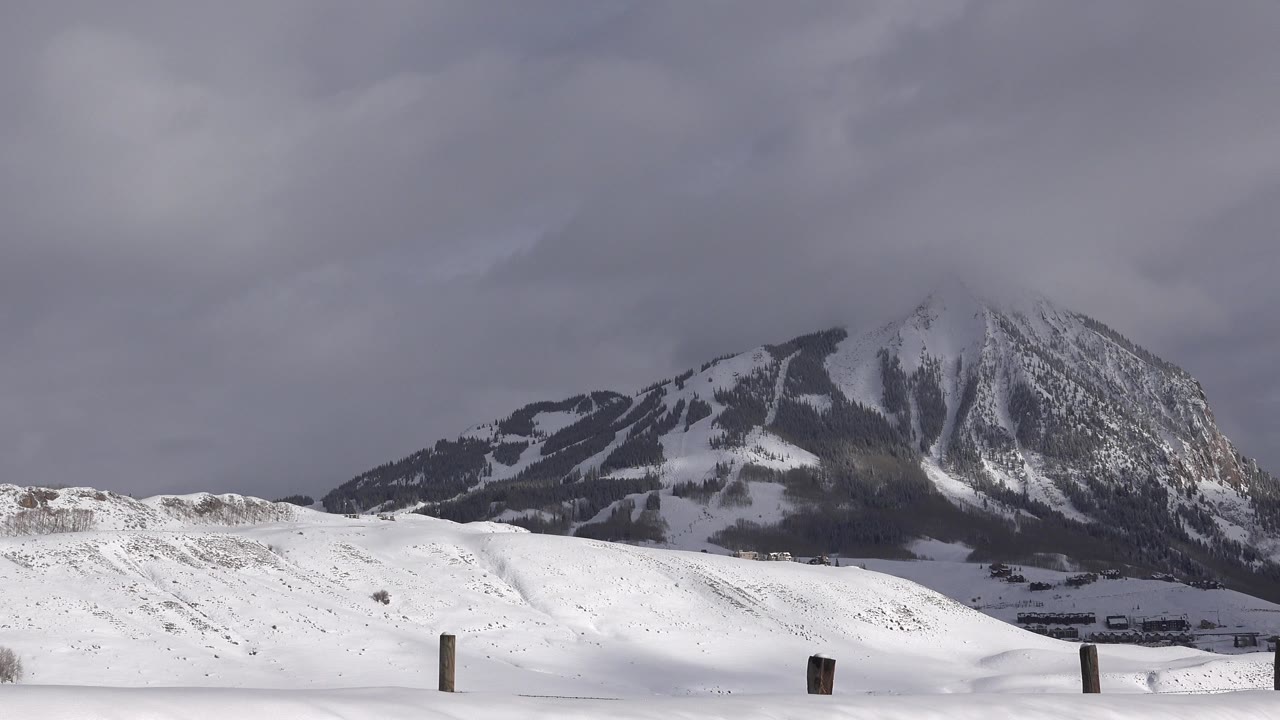 Wide view of snow-covered hills and a dark mountain ridge in Crested Butte, Colorado, with gray clouds over the peak and no visible people.