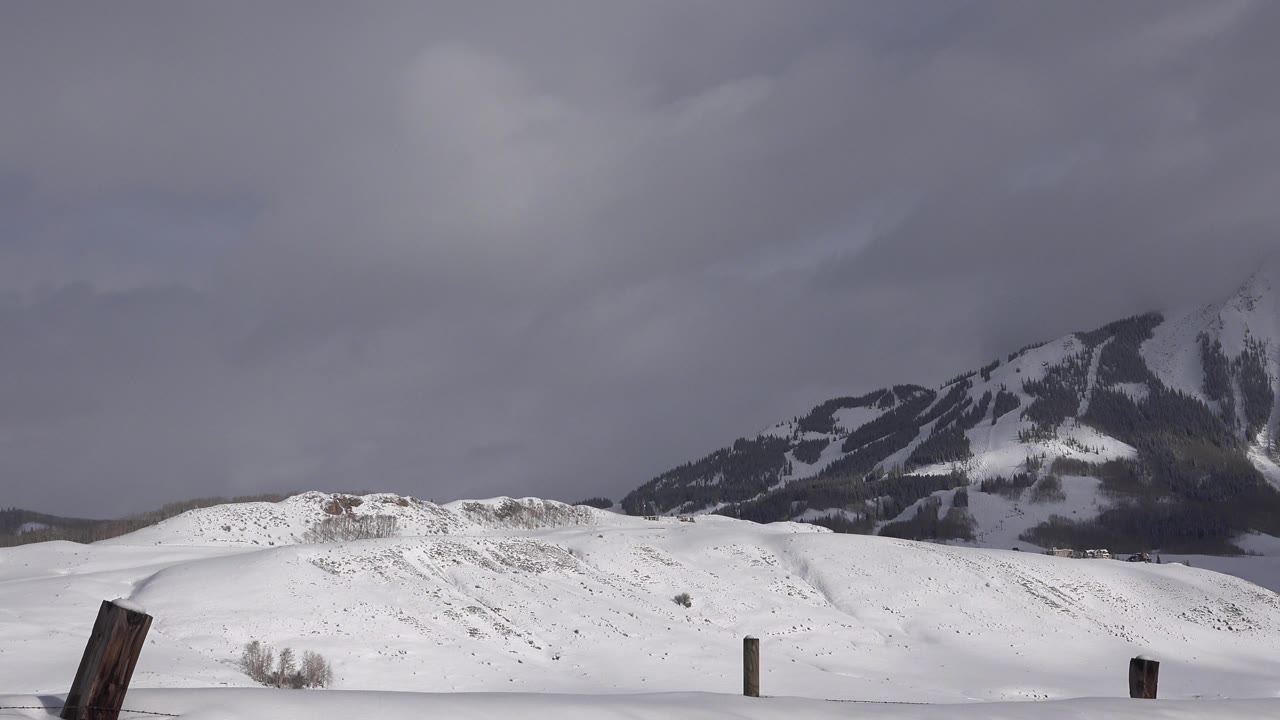 Opening view of snowy hills near Crested Butte, Colorado, with a dark mountain ridge behind them, gray cloud cover overhead, leaning wooden fence posts in the foreground, and no visible people.