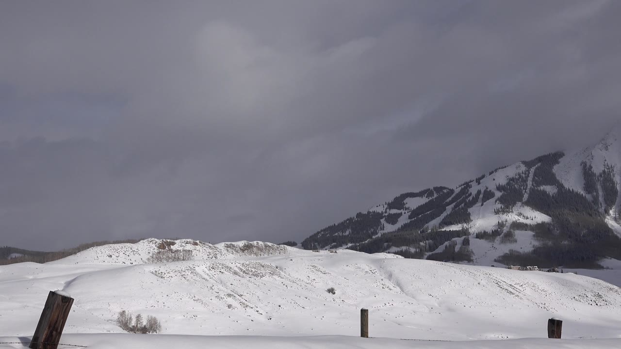 Snow-covered rolling ground in Crested Butte, Colorado, stretches toward dark mountain slopes partly softened by low gray clouds, with scattered fence posts and no visible people.
