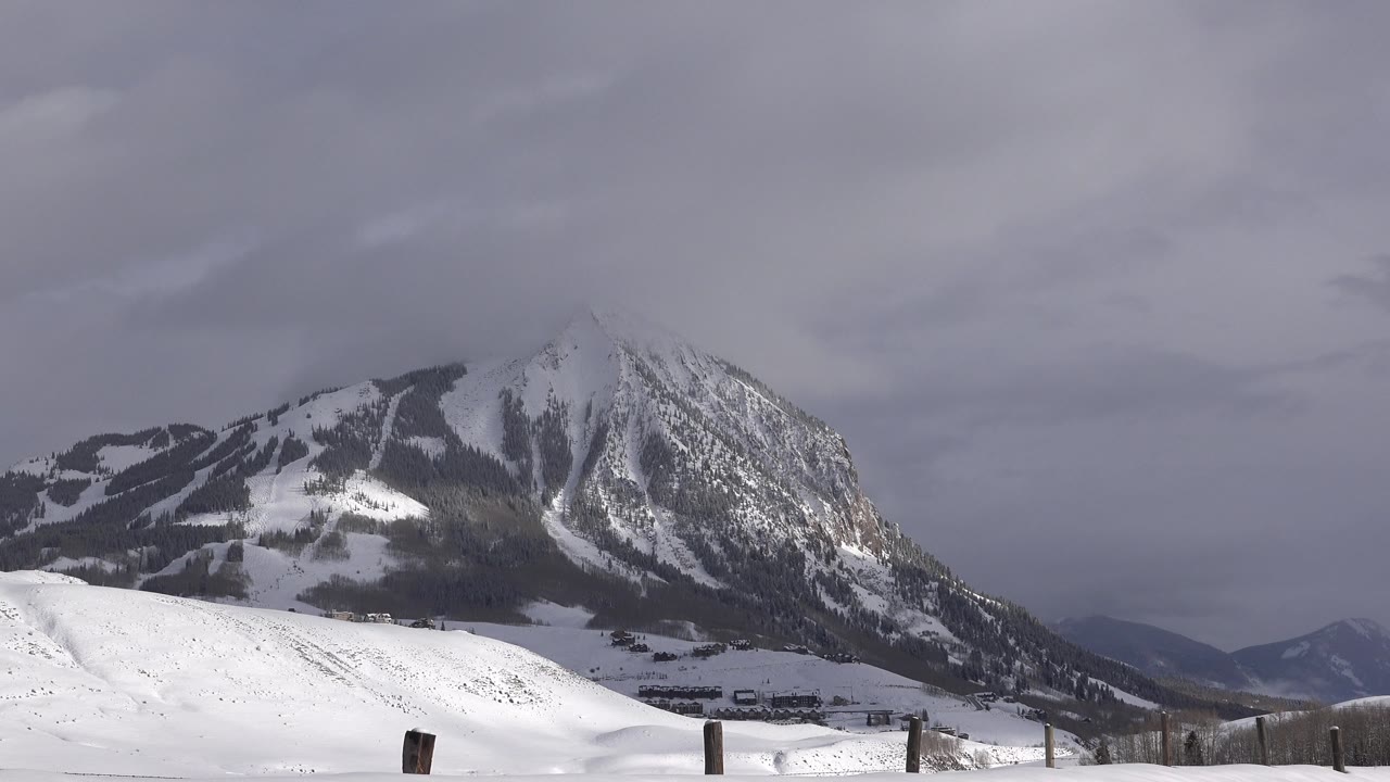 Mountain with snowy slopes, dark tree patches, and cloudy sky