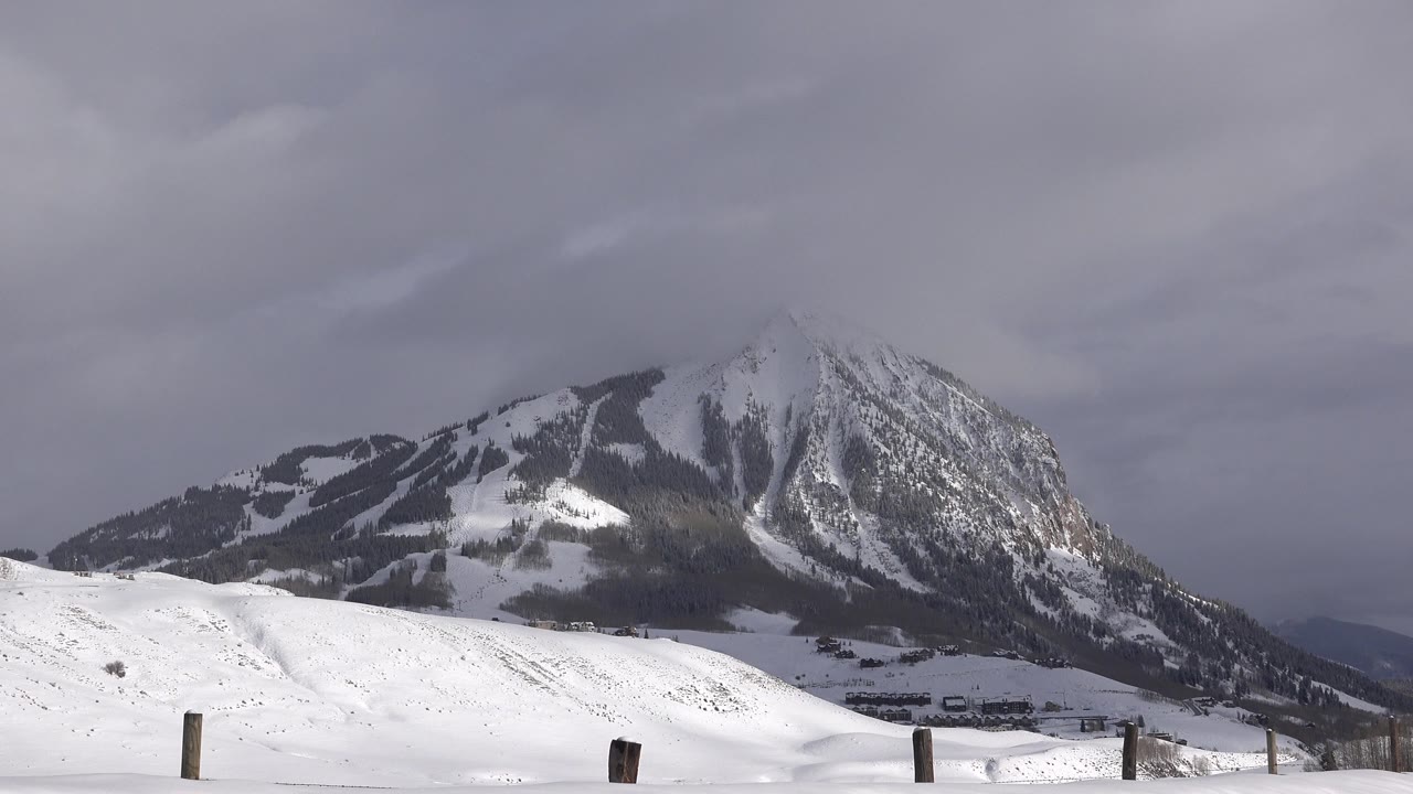 Snow-covered foreground with dark fence posts leads to a steep mountain under gray cloud cover in Crested Butte, Colorado, with no visible people.