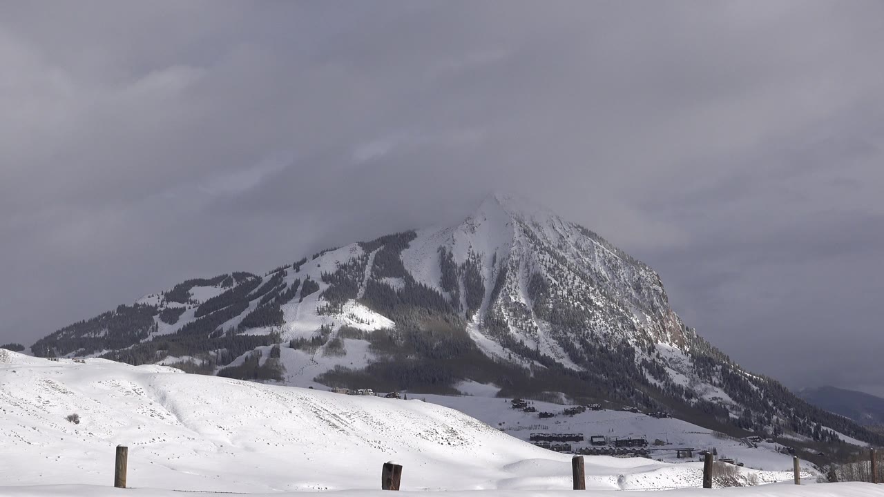 A snowy field with scattered dark fence posts sits below a broad mountain face and low gray clouds in Crested Butte, Colorado, with no visible people.