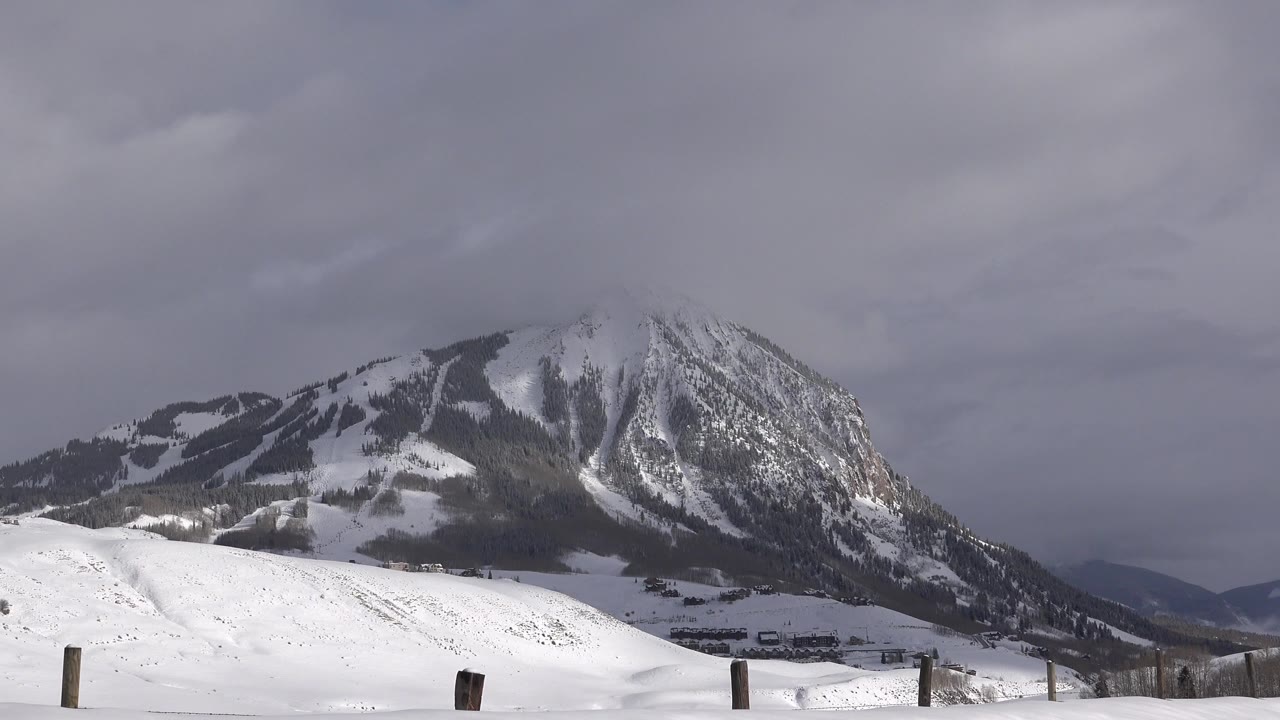 Wide Crested Butte, Colorado winter view with a snow-covered mountain, gray cloud cover, low fence posts, and no visible people at the opening moment.
