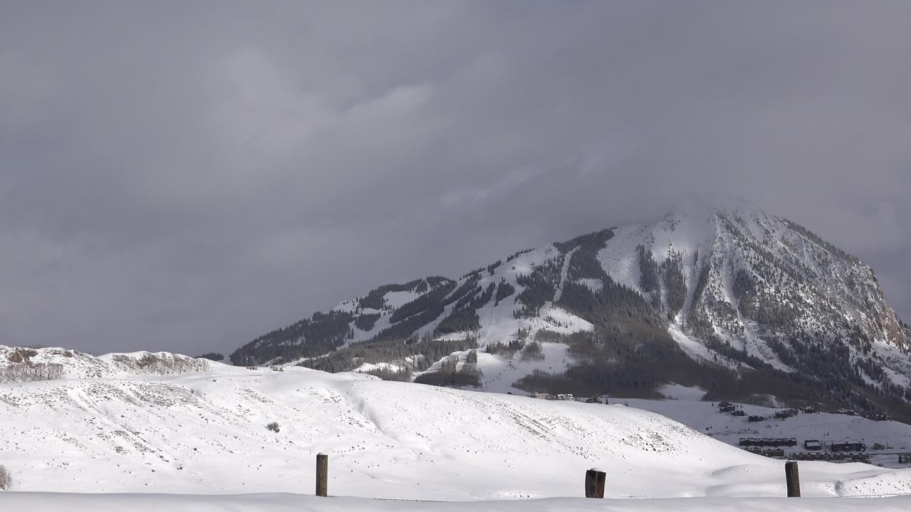 Opening frame of snow-covered hills in Crested Butte, Colorado, with a dark mountain behind them, gray clouds overhead, foreground fence posts, and no visible people.