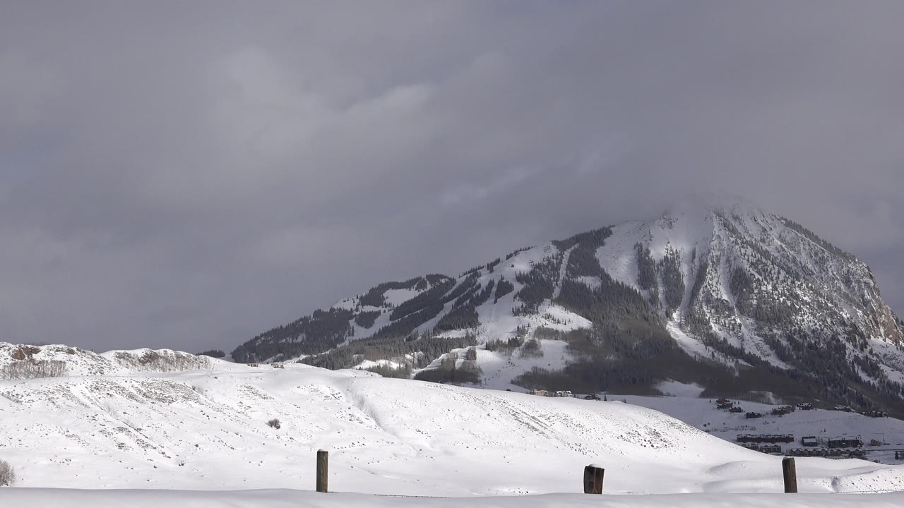 Second frame of the Crested Butte winter landscape showing a bright snow ridge, a darker mountain face, cloudy sky, scattered tree patches, and no visible people.
