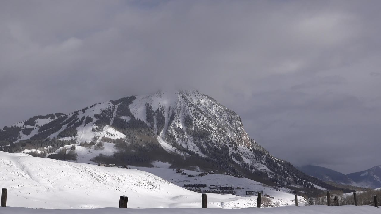 Snowy mountain under gray clouds with fence posts in the foreground.