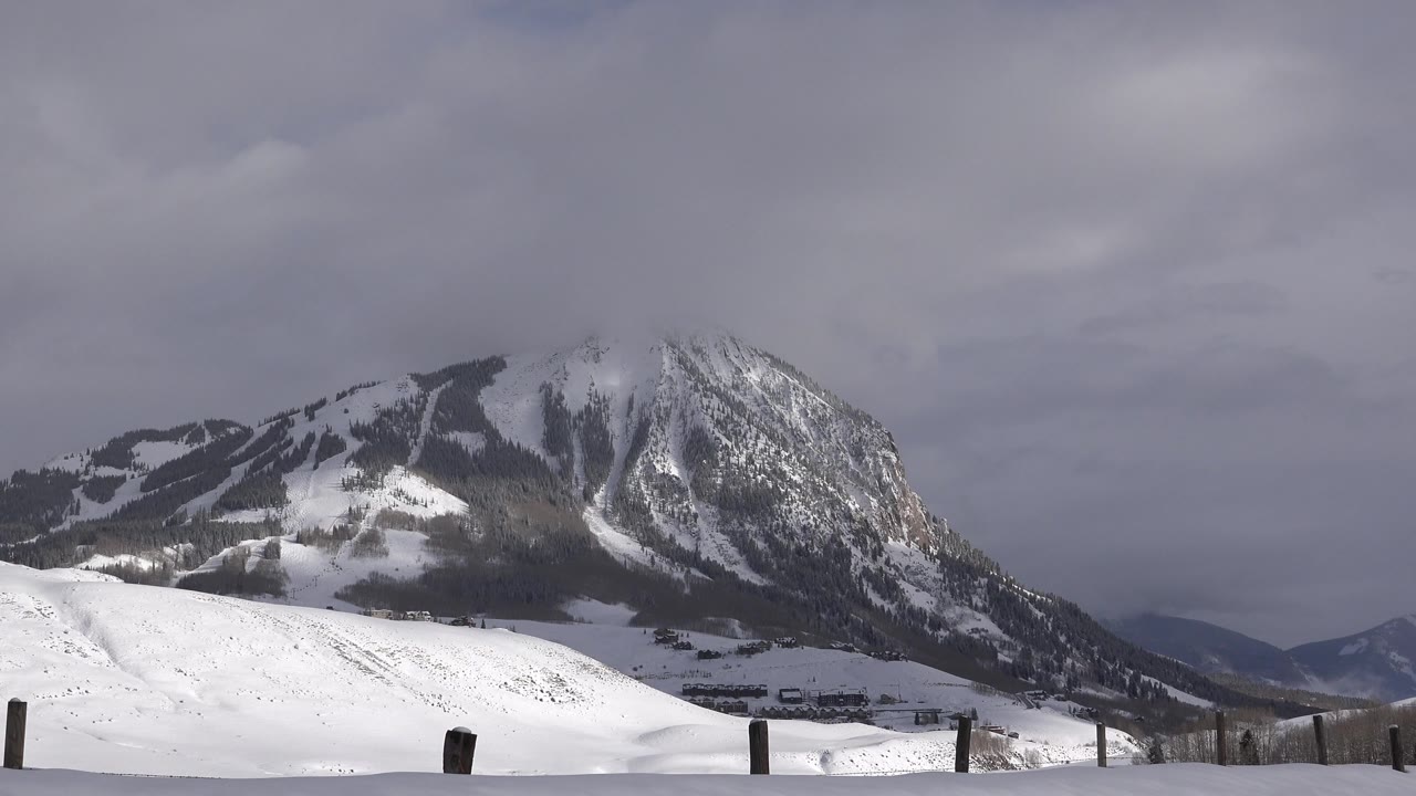 Early frame shows the Crested Butte mountain view slightly brighter across the foreground snow, with dark wooded bands on the slopes and no visible people.