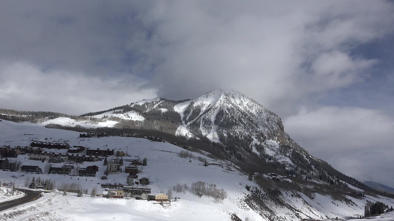 Wide Crested Butte, Colorado winter view with a snow-covered mountain rising behind village buildings, a curved road in the foreground, clouds overhead, and no visible people.