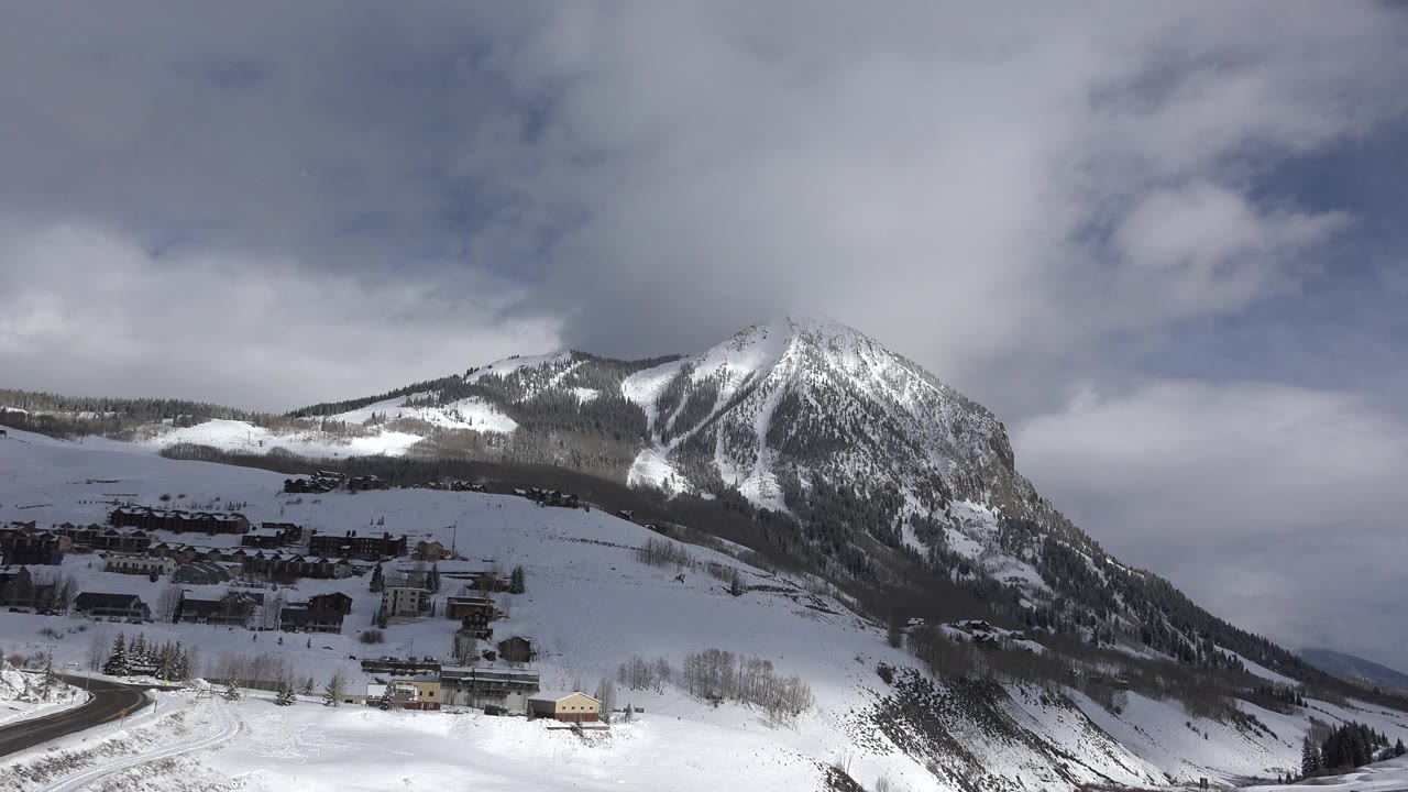 Crested Butte winter frame showing the mountain slightly farther left in the composition, with dark village rooftops, bright snowfields, a road edge, layered clouds, and no visible people.
