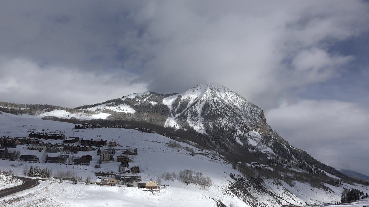 Winter village below a snow-striped mountain and cloudy sky.