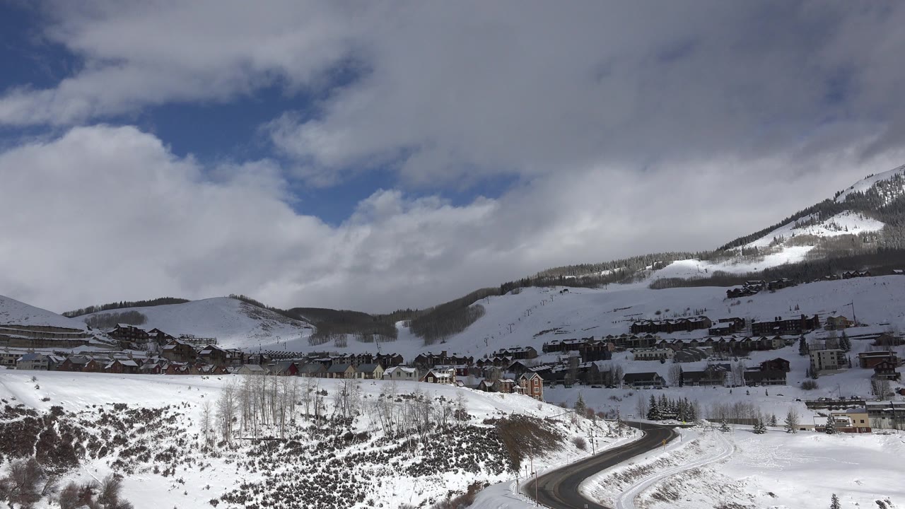 Wide winter view in Crested Butte, Colorado, showing snow-covered hills, a curving road, clustered buildings, mountains, and no visible people.