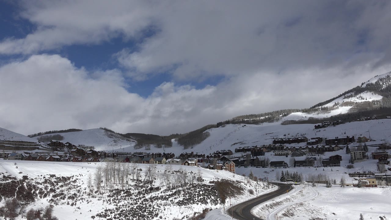 Crested Butte winter landscape with white hillsides, dark road curves, hillside buildings, mountain ridges, and no visible people.