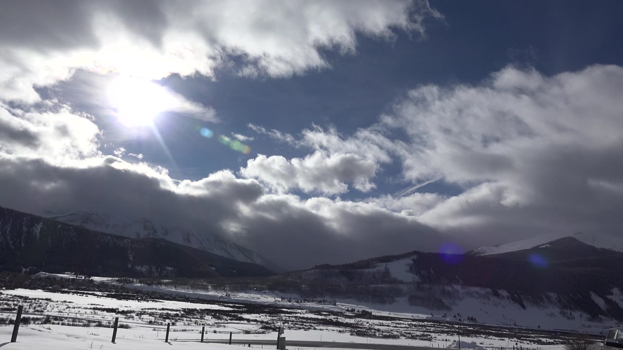 Bright sun breaks through clouds above snow-covered mountains in Crested Butte, Colorado, with fence posts in the foreground and no visible people.