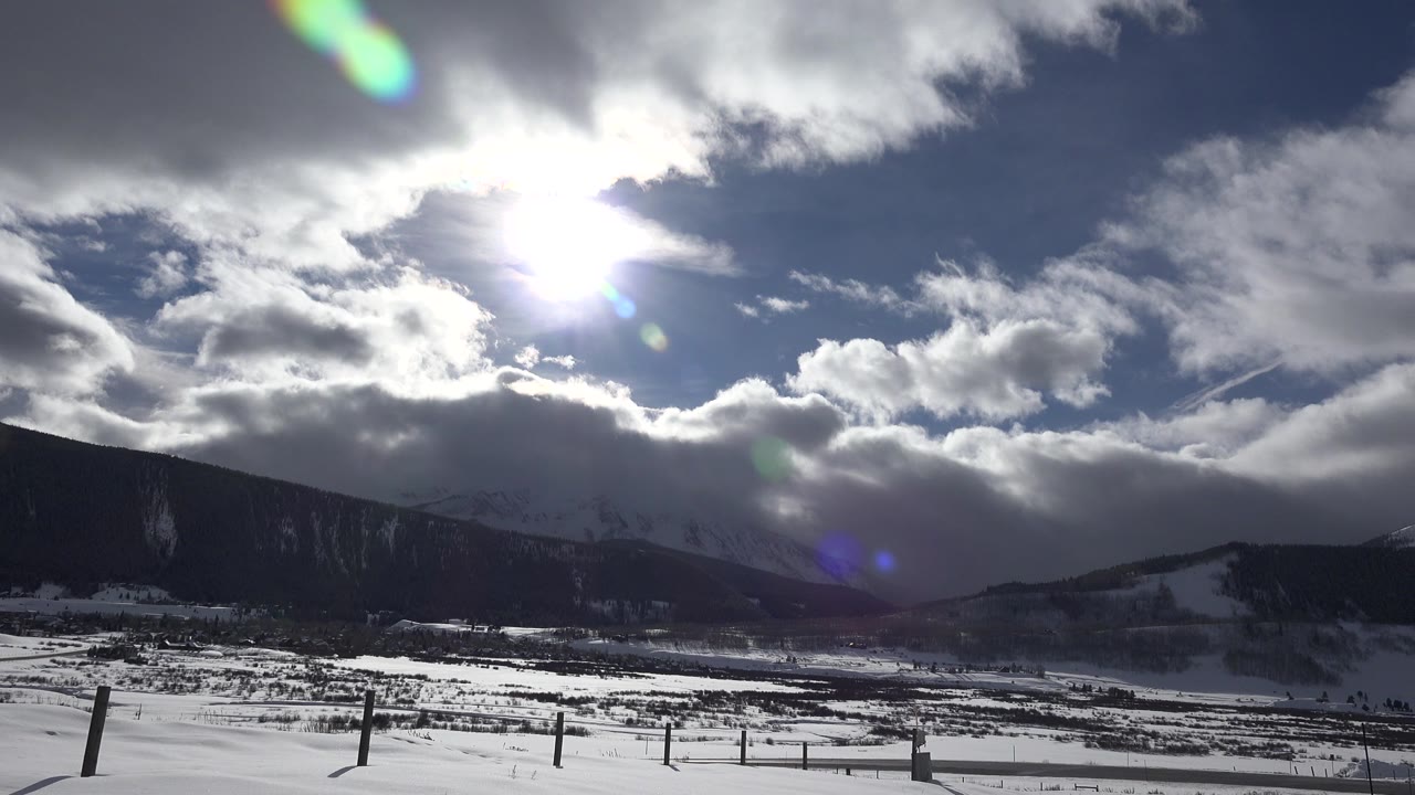 Bright sun and thick clouds hang over a snowy valley and dark mountain ridges in Crested Butte, Colorado, with fence posts in the foreground and no visible people.
