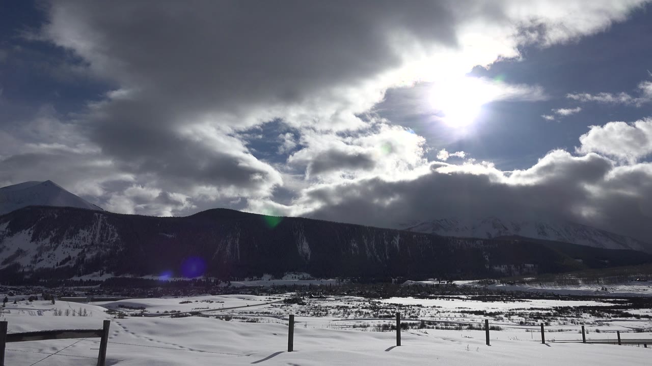 Snowy field, fence posts, dark mountain ridge, and bright sun behind clouds.