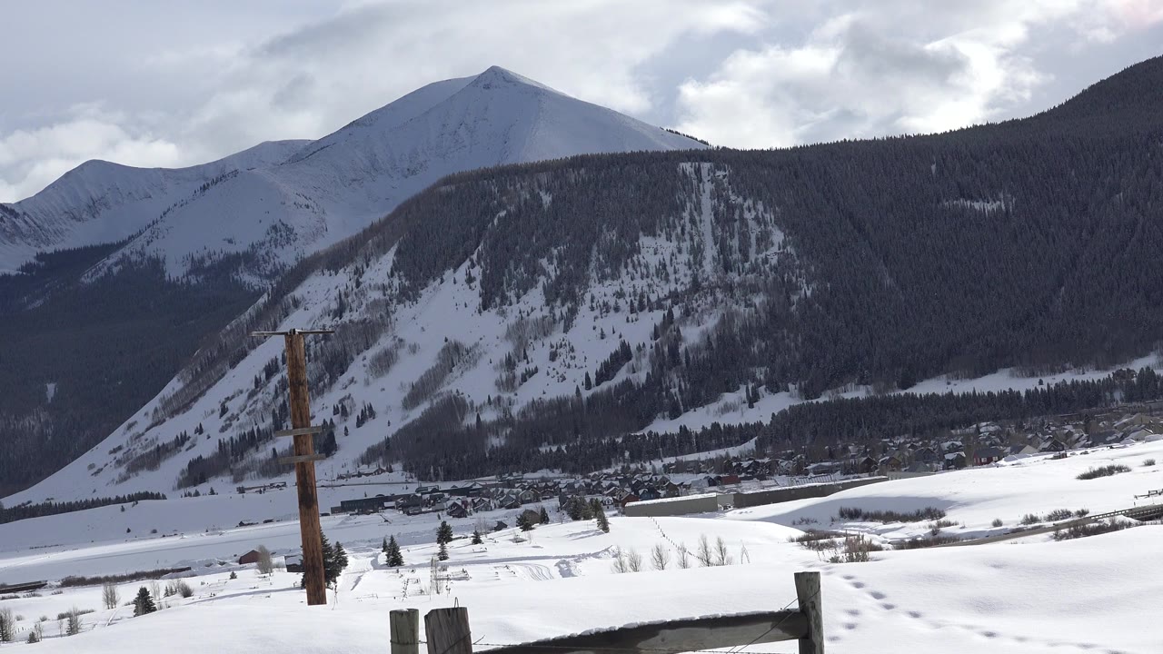 Wide Crested Butte, Colorado winter valley view with snowy peaks, dark forested slopes, foreground fence posts, a utility pole, road traces in the snow, and no visible people.