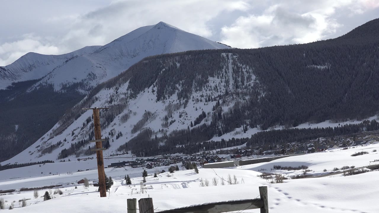 Crested Butte winter landscape with a pointed snowy peak, dark tree-covered ridge, small distant buildings in the valley, foreground snow, and no visible people.